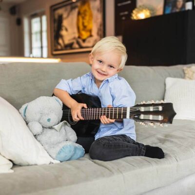 Familjefotografering hemma. Ett barn sittandes i soffa med nalle och gitarr.
