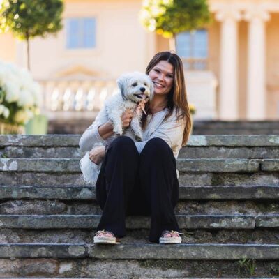 Porträtt av en liten vit hund i knät på sin matte. Foto taget på en husdjursfotografering utomhus vid Gunnebo slott.